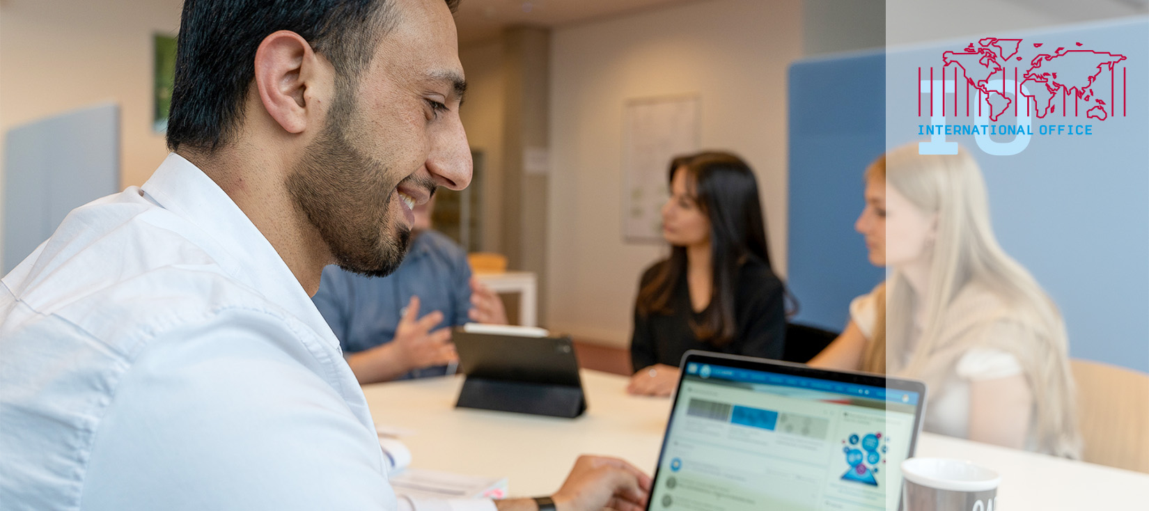 Student sitzt im Vordergrund und schaut auf sein Laptop. Im Hintergrund sitzen zwei Studentinnen im Gespräch.
