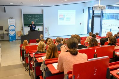 Zuschauer sitzen interessiert in einem Hörsaal. Am Rednerpult hält ein Mann eine Präsentation zu künstlicher Intelligenz.