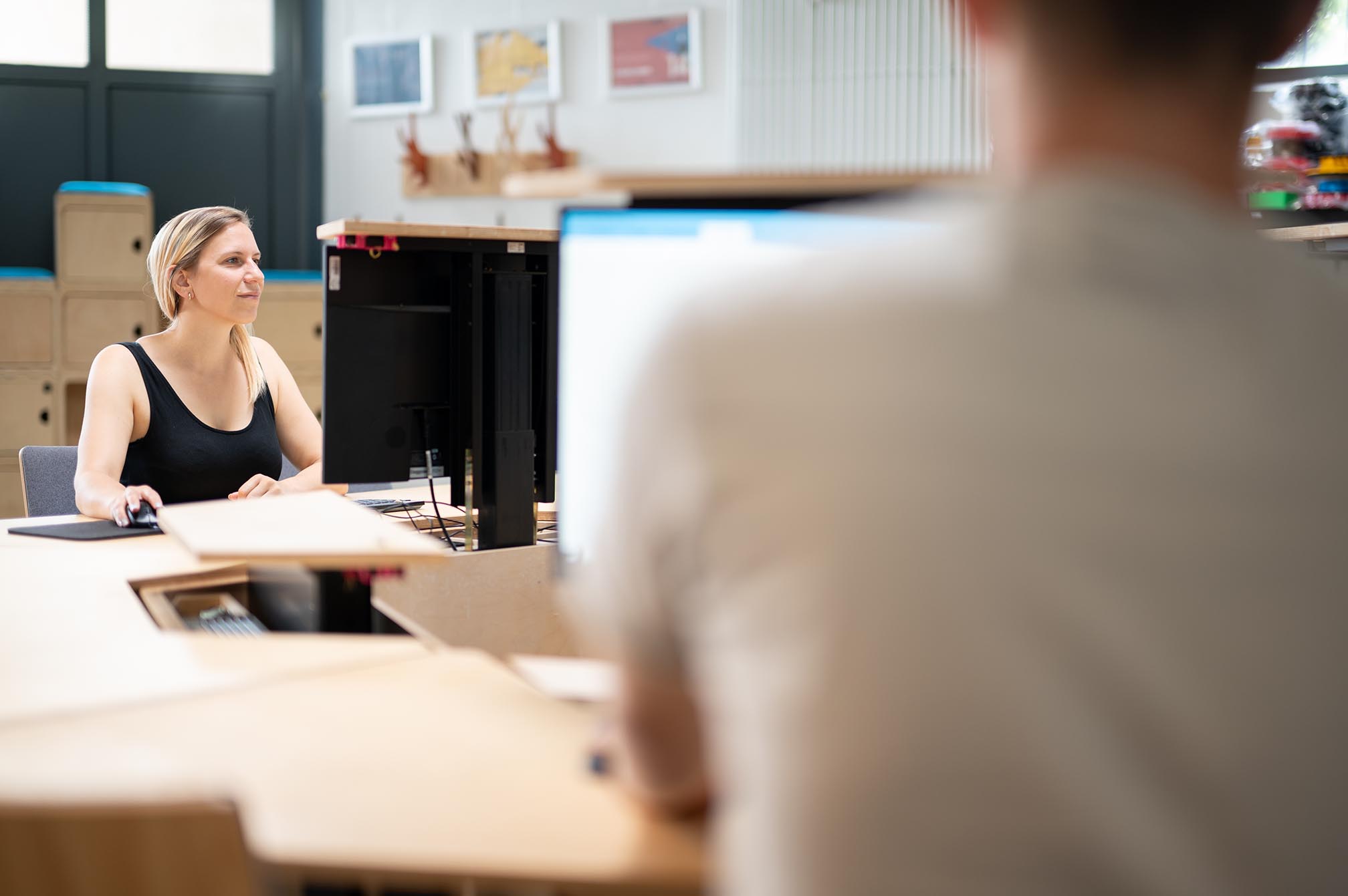 Eine Frau und ein Mann sitzen an verschiedenen Arbeitsplätzen in einem Büro und arbeiten an ihren Computern, wobei die Frau im Fokus des Bildes steht.