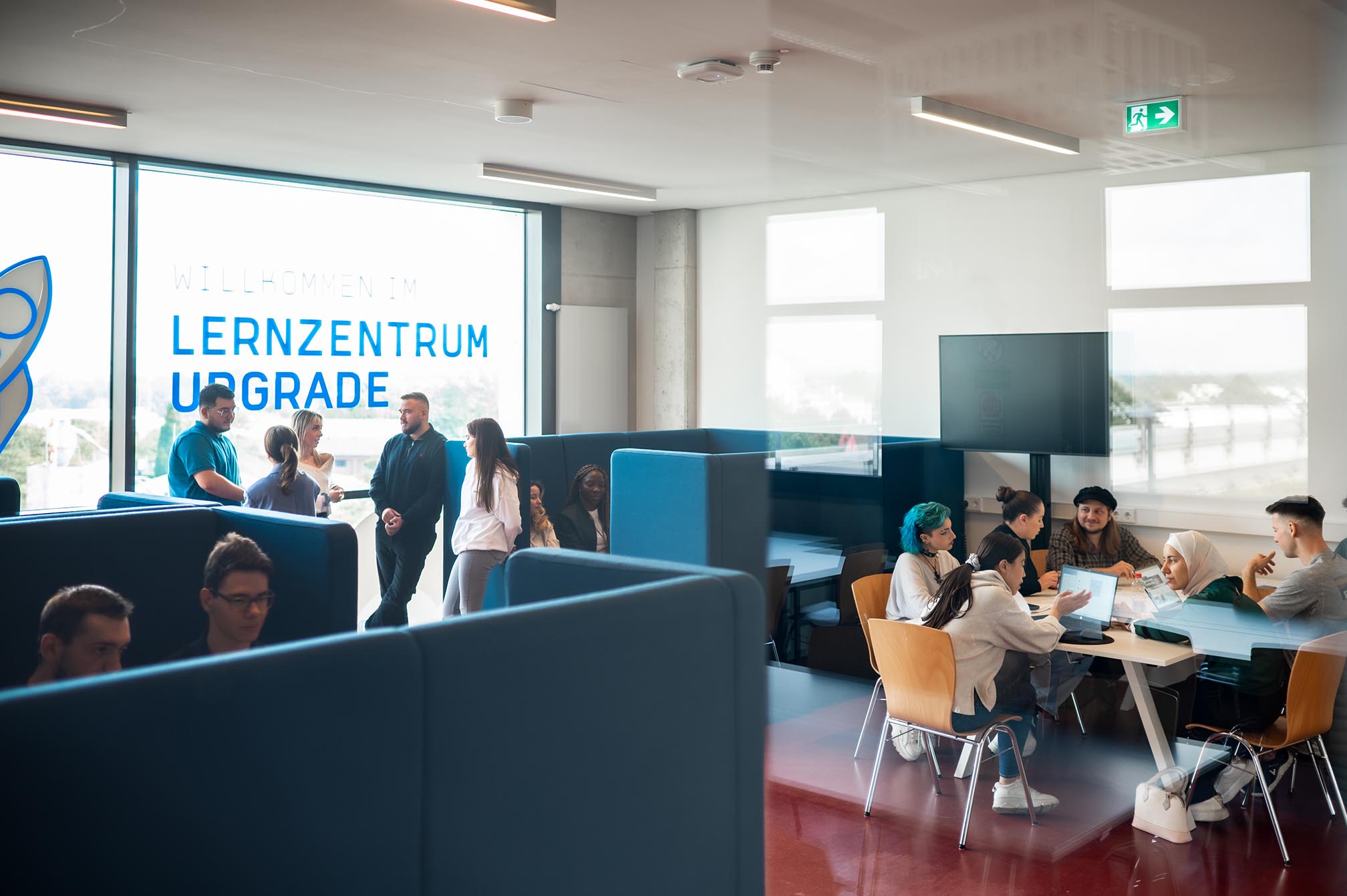 Studenten stehen und unterhalten sich an einem der Gebäude auf dem Campus Mülheim, neben den Lernplätzen, während andere in den Lernplätzen sitzen.