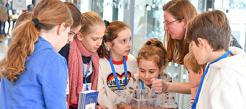 Auf dem Bild sieht man von links nach recht mehrere SchulKinder die mit einer erwachsenen Frau, die mit ihnen kommuniziert
